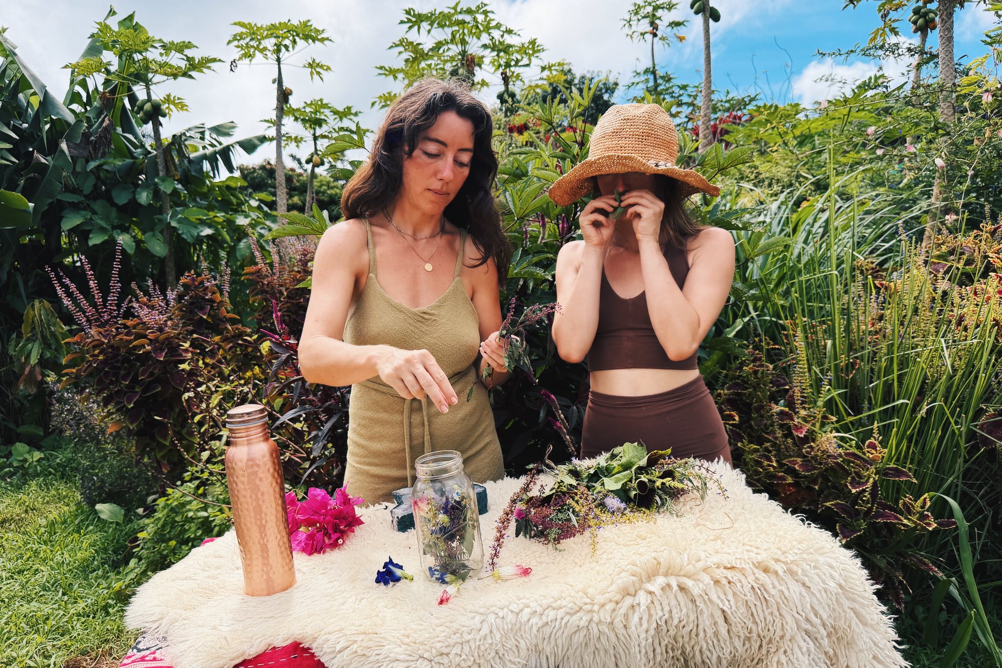 Two people at a garden table building a herbal infusion, cut herbs spread in front of them.