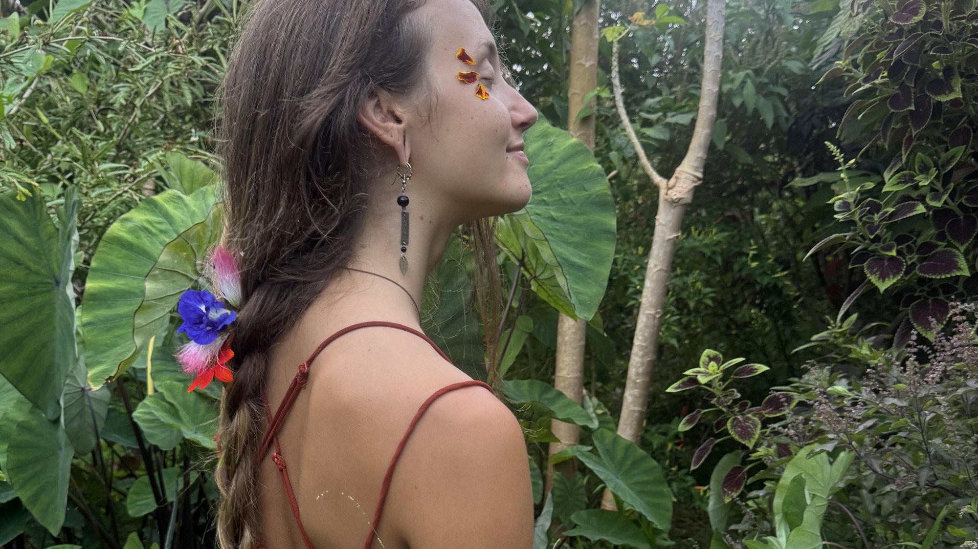 A person seated on the ground among kalo leaves and coleus, eyes closed.